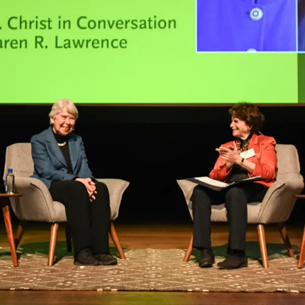 Two people sit in chairs on a stage. A large projection screen reads “Carol T. Christ in Conversation with Karen R. Lawrence.”