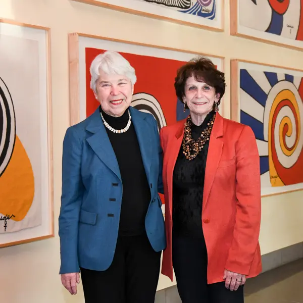 Two people stand together in front of a wall of art in primary colors.
